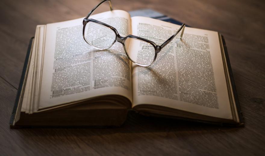 Een op een bruine tafel opengeslagen oud boek, met daarop een opengeklapte leesbril in grijs montuur.