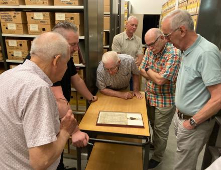 Vrijwilligers van het politiemuseum bekijken in het depot van het Gemeentearchief een bijzonder, ingelijst, archiefstuk. Op de achtergrond zijn stellingkasten met archiefdozen te zien.